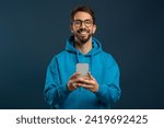 Portrait Of Smiling Handsome Young Man With Smartphone In Hands, Happy Millennial Guy Wearing Hoodie And Eyeglasses Using Mobile Phone While Standing Against Blue Studio Background, Copy Space