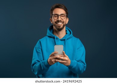 Portrait Of Smiling Handsome Young Man With Smartphone In Hands, Happy Millennial Guy Wearing Hoodie And Eyeglasses Using Mobile Phone While Standing Against Blue Studio Background, Copy Space - Powered by Shutterstock