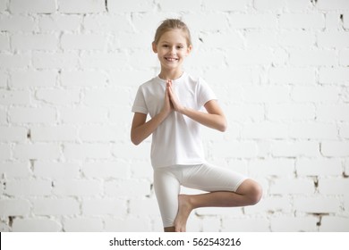 Portrait Of Smiling Happy Girl Child Practicing Yoga, Standing In Vrksasana Exercise, Tree Pose With Namaste, Working Out Wearing Sportswear, T-shirt, Pants, Indoor, White Studio Background 