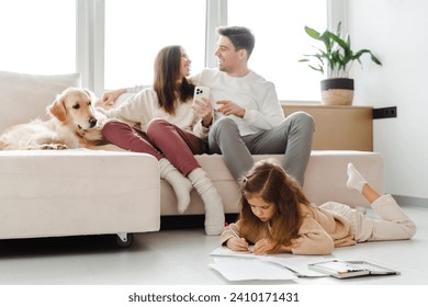 Portrait of smiling happy mother and father, woman holding mobile phone, dog lying on sofa near them, little daughter sitting on floor drawing, in cozy home. Concept of parenthood, online shopping - Powered by Shutterstock