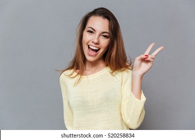 Portrait of a smiling happy woman showing victory sign and looking at camera isolated on the gray background