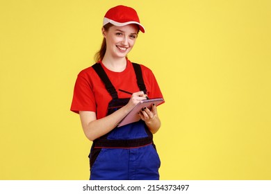 Portrait of smiling happy worker woman writing in paper notebook, making to do list, writes down orders, wearing overalls and red cap. Indoor studio shot isolated on yellow background. - Powered by Shutterstock