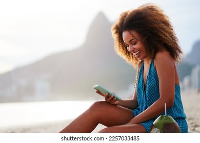 portrait smiling happy young brazilian woman sitting on the beach holding and looking at cell phone in Ipanema - Powered by Shutterstock