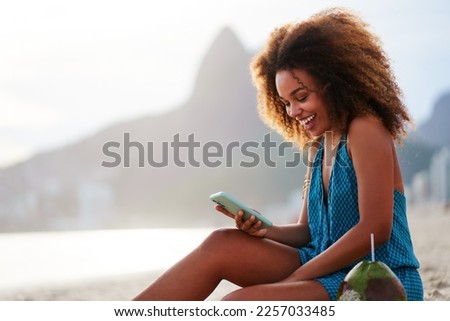 portrait smiling happy young brazilian woman sitting on the beach holding and looking at cell phone in Ipanema