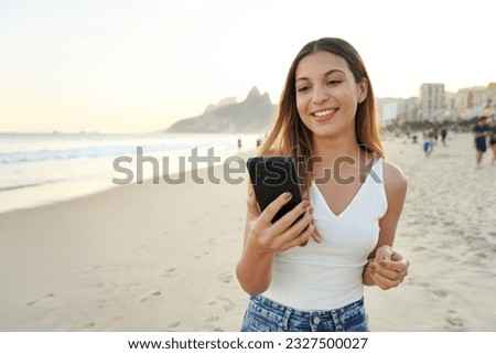 Portrait of smiling happy young Brazilian woman on Ipanema beach holding and watching her smartphone, Rio de Janeiro, Brazil