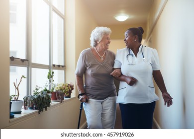 Portrait Of Smiling Healthcare Worker Walking And Talking With Senior Woman. Happy Elder Woman Gets Help From Nurse For A Walk Through Nursing Home.