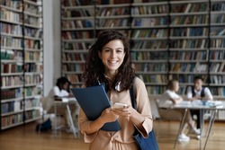 Portrait Of Smiling Hispanic Female College Student With Headphones In Neck Holding Folder And Cellphone In Hands, Using Different Modern Tech Gadgets In Public Library, Browsing Internet, Web Surfing