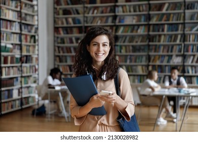 Portrait Of Smiling Hispanic Female College Student With Headphones In Neck Holding Folder And Cellphone In Hands, Using Different Modern Tech Gadgets In Public Library, Browsing Internet, Web Surfing