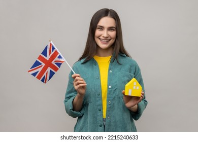 Portrait Of Smiling Holding Great Britain Flag And Paper House, Dreaming To Buy Accommodation In England, Wearing Casual Style Jacket. Indoor Studio Shot Isolated On Gray Background.