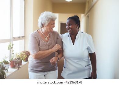 Portrait of smiling home caregiver and senior woman walking together through a corridor. Healthcare worker taking care of elderly woman. - Shutterstock ID 421368178