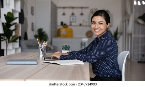 Portrait smiling Indian businesswoman using laptop at home office workplace, typing on keyboard, happy successful confident young woman freelancer looking at camera, working or studying online