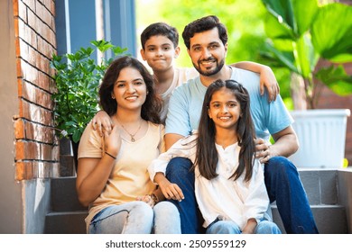 Portrait of Smiling Indian family of four, young parents and two kids, sitting joyfully on the front steps of their home, sharing a warm and happy moment together, celebrating love, family bond - Powered by Shutterstock