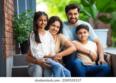 Portrait of Smiling Indian family of four, young parents and two kids, sitting joyfully on the front steps of their home, sharing a warm and happy moment together, celebrating love, family bond - Powered by Shutterstock