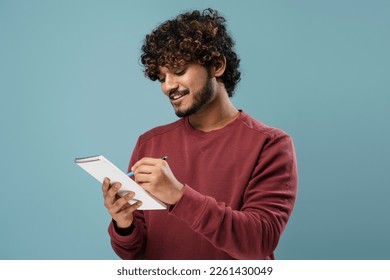 Portrait of smiling Indian man taking notes in notepad isolated on blue background. Smart asian university student studying, learning language, exam preparation. Education concept