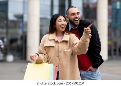 Portrait Of Smiling International Couple Holding Shopper Bags, Pointing At Store Window, Walking Near Shopping Center. Millennial Diverse Family Feeling Happy And Excited Next To Mall