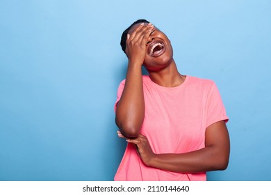 Portrait of smiling joyful african american young woman laughting at camera while standing in studio with blue background. Teenager making happy facial expressions. Photography concept