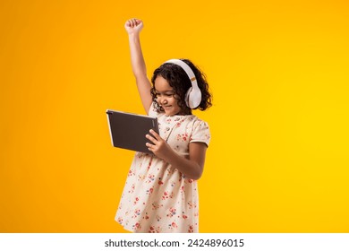 Portrait of smiling kid girl with headphones using tablet and showing winner gesture. Lifestyle, leasure and gadget addiction concept - Powered by Shutterstock