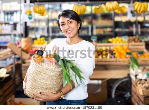 Portrait of smiling\
latin american woman with wicker basket with fresh grocery,\
enjoying shopping in\
store