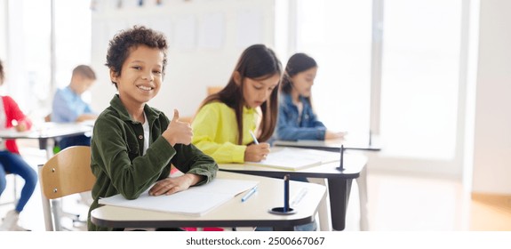 Portrait of smiling Latin schoolboy showing thumbs up and smiling, sitting at desk in his classroom with classmates on background. - Powered by Shutterstock