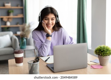 Portrait Of Smiling Laughing Young Woman Sitting At Table, Wearing Wireless Headset And Using Laptop. Young Female Watching Video, Movie Or Webinar, Having Video Call With Friends Or Family