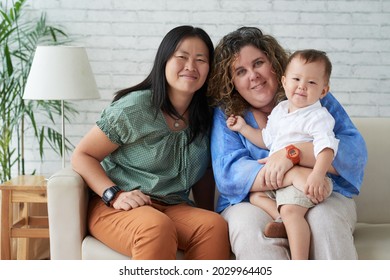 Portrait Of Smiling Lesbian Couple Sitting On Sofa In Living Room With Their Little Son On Laps