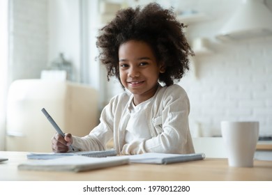 Portrait of smiling little African American girl child feel excited study learn at home kitchen. Happy small 8s biracial ethnic kid do homework assignment in notebook. Education, diversity concept. - Shutterstock ID 1978012280