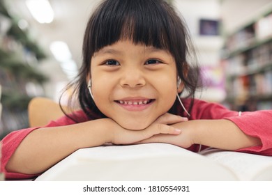 Portrait Of A Smiling Little Asian Girl. Children Smile And Looking Camera. Happy Asia Kid In Pink Shirt And Reading Book  And Listen Music At Library. Education People Concept