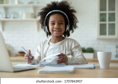 Portrait of smiling little biracial girl in headphones do homework study online in kitchen, happy small African American child in earphones have online web class or lesson using laptop at home