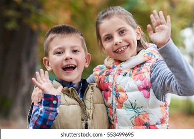 Portrait Of A Smiling Little Brother And Sister Waving Hello While Enjoying A Day Outside Together In A Park In The Autumn