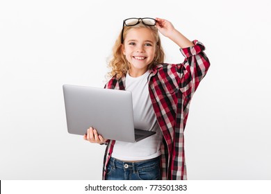 Portrait of a smiling little girl in eyeglasses holding laptop computer while standing and looking at camera isolated over white background