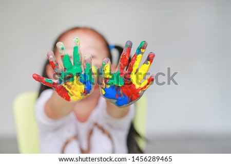 Portrait of smiling little girl looking through her colorful hands and cheek painted in kids room. Focus at baby hands.