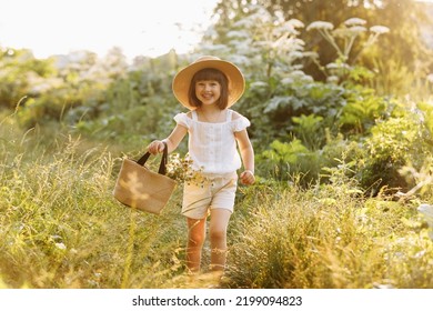 Portrait of smiling little girl walks in the rays of a sunset in a flowering meadow, enjoying the summer, warmth, flowers, freedom. Child with straw hat and bag is having fun outdoors.