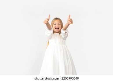 Portrait of smiling little girl wearing white dress showing thumbs up, like gesture, looking at camera demonstrating her satisfaction. Indoor studio shot isolated on pink background
