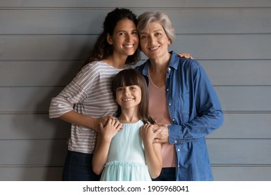Portrait smiling little girl with young mother and mature grandmother standing on grey wooden wall background, looking at camera, three generations of women posing for family photo, multigenerational