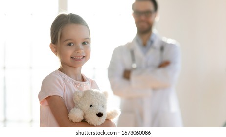 Portrait Of Smiling Little Patient Holding Favorite Toy With Pleasant Male Doctor On Background. Happy Small Girl Posing For Photo With Male General Practitioner, Preschooler Visiting Doc In Clinic.