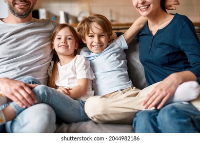 Portrait of smiling little teen kids sit on sofa relax at home with young Caucasian parents. Happy small teenage children siblings rest with mother and father, rest together. Parenthood concept.