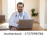 Portrait of smiling male Caucasian doctor wearing whit medical uniform sit at desk in hospital look ta camera posing, happy young man GP or physician work on modern laptop in private clinic