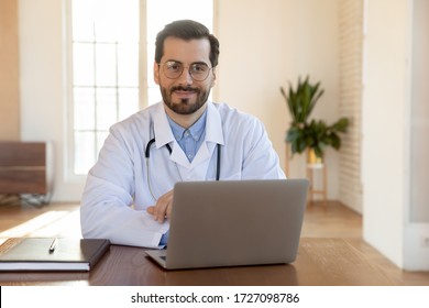 Portrait Of Smiling Male Caucasian Doctor Wearing Whit Medical Uniform Sit At Desk In Hospital Look Ta Camera Posing, Happy Young Man GP Or Physician Work On Modern Laptop In Private Clinic