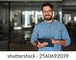 Portrait of smiling male company worker employee, project manager or entrepreneur in office. Happy young professional Latin business man looking at camera standing at work using tablet.