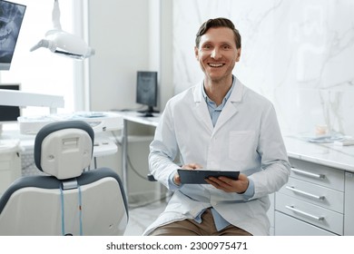 Portrait of smiling male dentist looking at camera in dental clinic interior and holding clipboard, copy space - Powered by Shutterstock