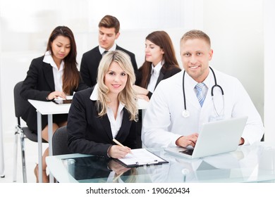 Portrait Of Smiling Male Doctor And Businesswoman With Laptop Sitting At Desk In Office