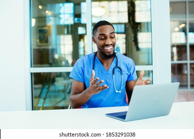 Portrait of a smiling male doctor wearing blue scrubs uniform using laptop to talk to patient online