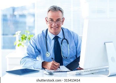 Portrait Of Smiling Male Doctor Writing Data At Desk In Hospital