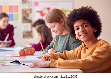 Portrait Of Smiling Male Elementary School Student Working At Desk In Classroom With Teacher - Powered by Shutterstock