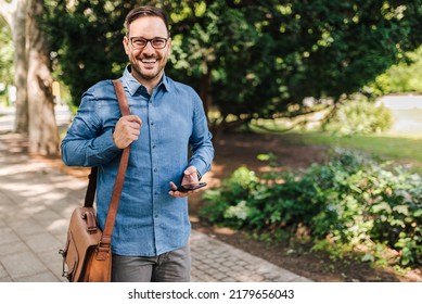 Portrait Of Smiling Male Executive Walking On His Way To Work. Young Professional Commuter With Smart Phone Is Carrying Laptop Bag. He Is Wearing Formals In The The City Park.