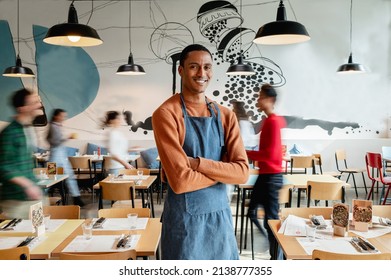 Portrait Of Smiling Male Owner Or Waiter In Cafe With Unrecognizable Blurred Employes Moving.