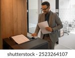 Portrait of smiling male printer in eyeglasses holding documents and operating computer printer in office