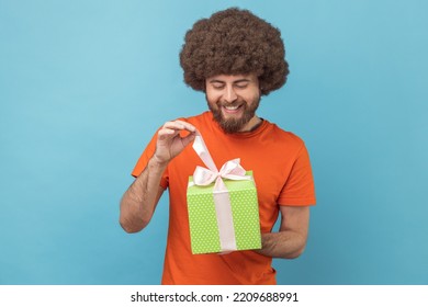 Portrait Of Smiling Man With Afro Hairstyle Wearing Orange T-shirt Unwrapping Birthday Present For Celebrating Holiday, Pulling Ribbon, Looking At Gift. Indoor Studio Shot Isolated On Blue Background.