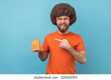 Portrait of smiling man with Afro hairstyle wearing orange T-shirt and funny eyeglasses holding pointing at paper house, helping with rent. Indoor studio shot isolated on blue background. - Shutterstock ID 2250245591