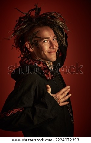 Portrait of a smiling man in black ethnic clothing with dreadlocks on his head and tattoos. Dark red background. 